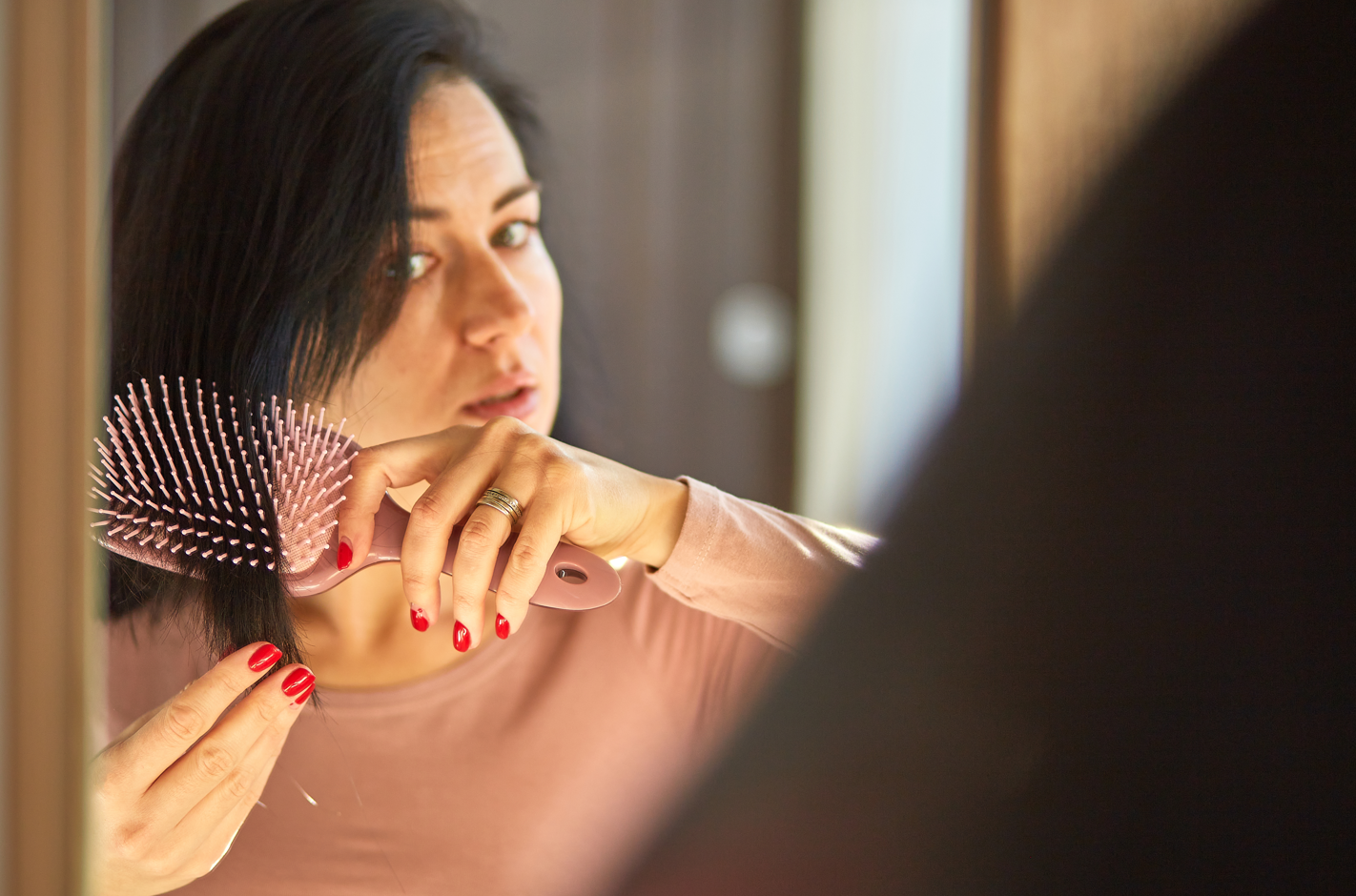 A woman with black and thinning hair combs her hair using a hair brush while looking into a mirror.