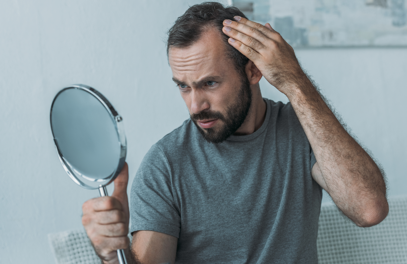 A bearded man with thinning hair and receding hairline looks at a handheld mirror with worry. Concept of thinning hair, lack of confidence, hair loss, self esteem.