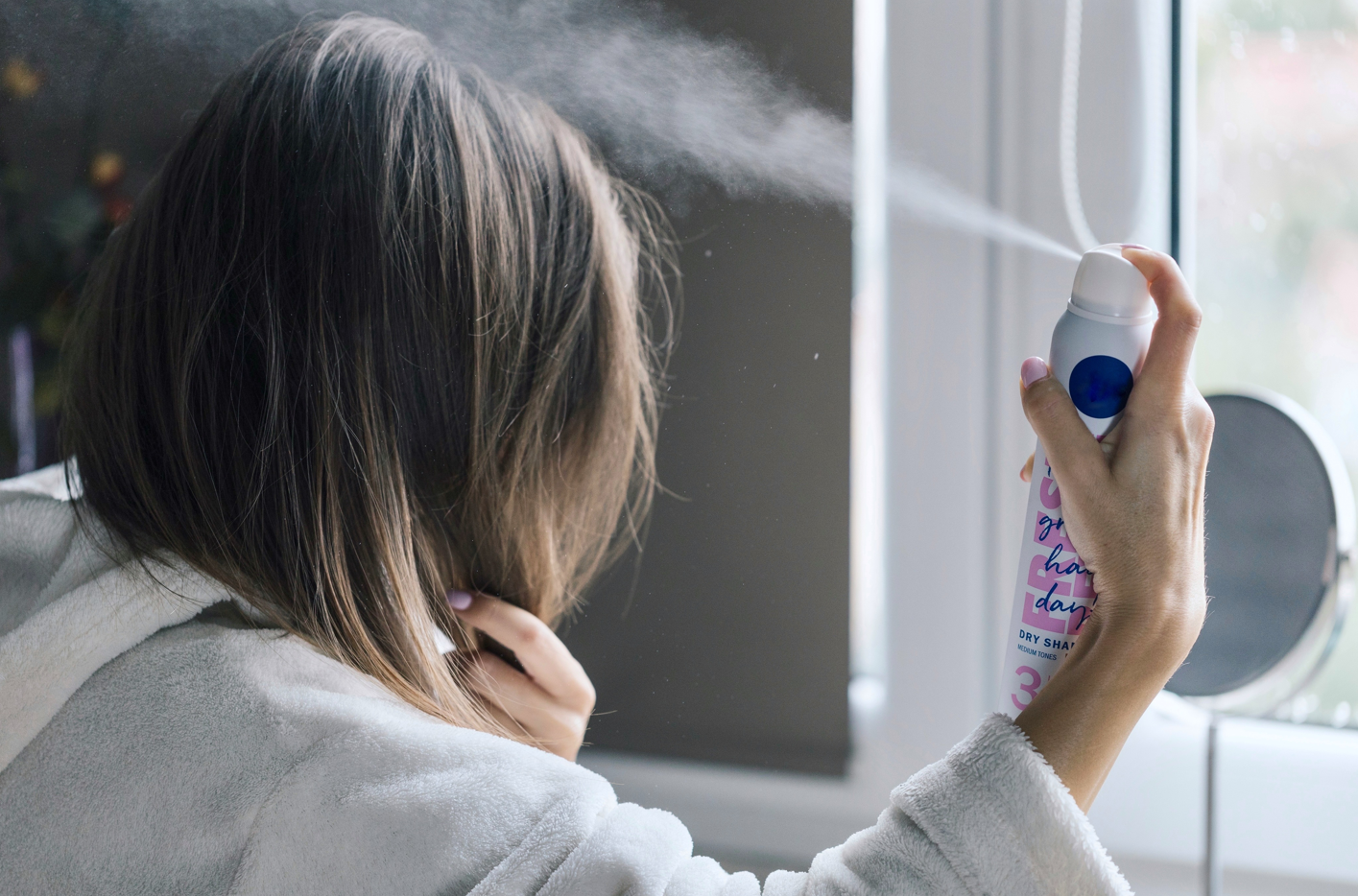 A woman with brown hair spraying dry shampoo on her hair. How to choose the best dry shampoo, right dry shampoo for your hair.